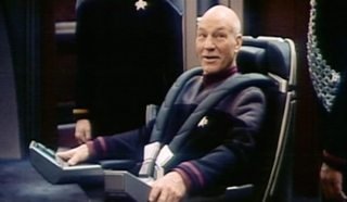 Picard sits in the captain's chair looking up at Worf who stands besides him; he has a black thick harness on to hold him in the seat