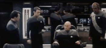 Gif showing Picard sitting in a chair surrounded by a few people, he presses a button on the chair's console and a seat belt harness shoots out over his lap and shoulders