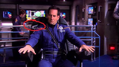 Riker sits at the chair looking down as he settles into it, his arms on the arm rests. A circle highlights a silver outlined hole in the back likely to be used for a seat belt/harness