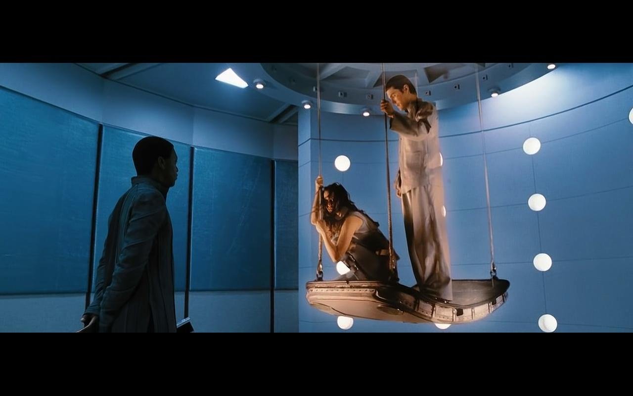 A dark blue tinted room. A lone figure faces obliquely away from the camera. They are viewing two holographic figures, tinted beige. One of the figures is female in appearance and crouching the other one male, standing. Both are holding vertical bars of the platform they are standing on